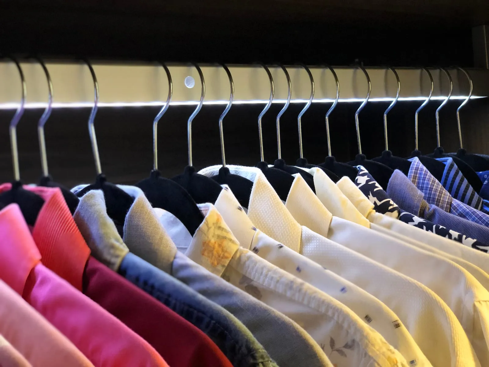 Camisas colgadas en perchas iguales y ordenadas por color, del rosa al amarillo y al azul, bajo una barra con luz.