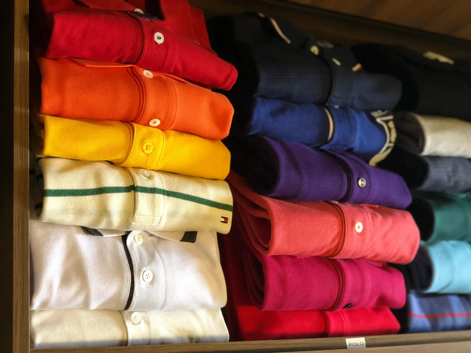 Cajón de polos doblados en pilas y ordenados por color: rojo, naranja, amarillo, azul, morado y rosa.