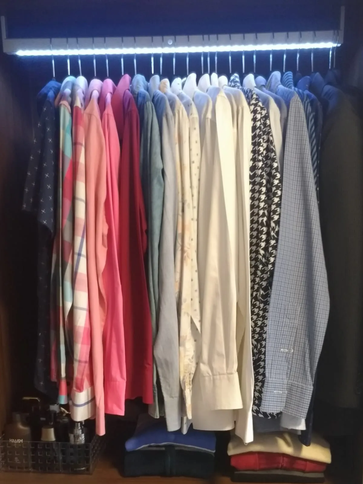 Barra de vestidor iluminada con blusas y camisas colgadas en perchas iguales, agrupadas por color.