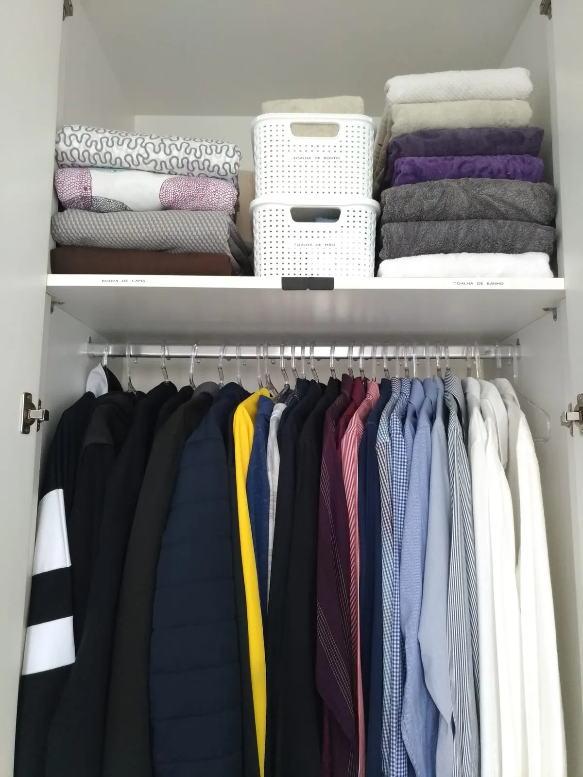 Armario con toallas y ropa doblada en cestas blancas arriba y chaquetas y camisas colgadas por color abajo.