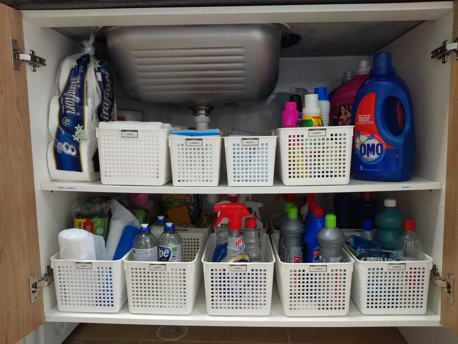 Mueble bajo el fregadero con dos baldas de cestas blancas que separan detergentes y productos de limpieza.